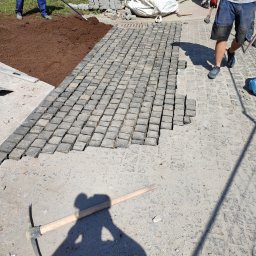 TEKA BRUK SPÓŁKA Z OGRANICZONĄ ODPOWIEDZIALNOŚCIĄ - Fragment nawierzchni z kostki granitowej w trakcie układania; widoczne przygotowane podłoże, ułożona część nawierzchni, narzędzia i pracownicy.