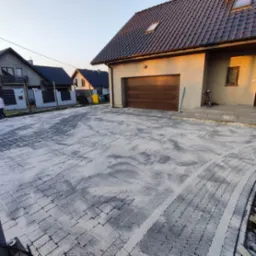 Nowo ułożona, szara kostka brukowa na podjeździe do domu jednorodzinnego, zasypana piaskiem, z widocznymi śladami po oponach samochodu.