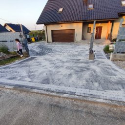 TEKA BRUK SPÓŁKA Z OGRANICZONĄ ODPOWIEDZIALNOŚCIĄ - Nowo ułożona, szara kostka brukowa na podjeździe do domu jednorodzinnego, z widocznymi śladami piasku stabilizującego i przechodząca obok bramy wjazdowej, po której przechodzi dziewczynka.