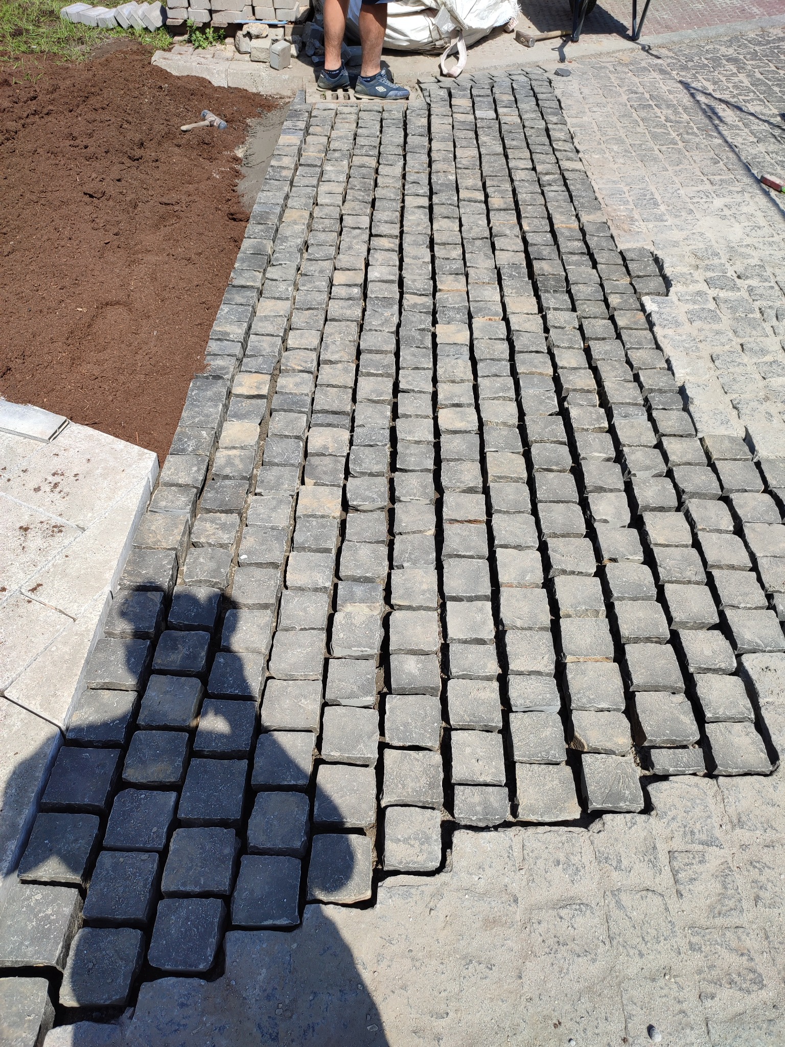 Fragment układanej nawierzchni z ciemnej kostki granitowej, widoczne postępy prac i cień osoby wykonującej.