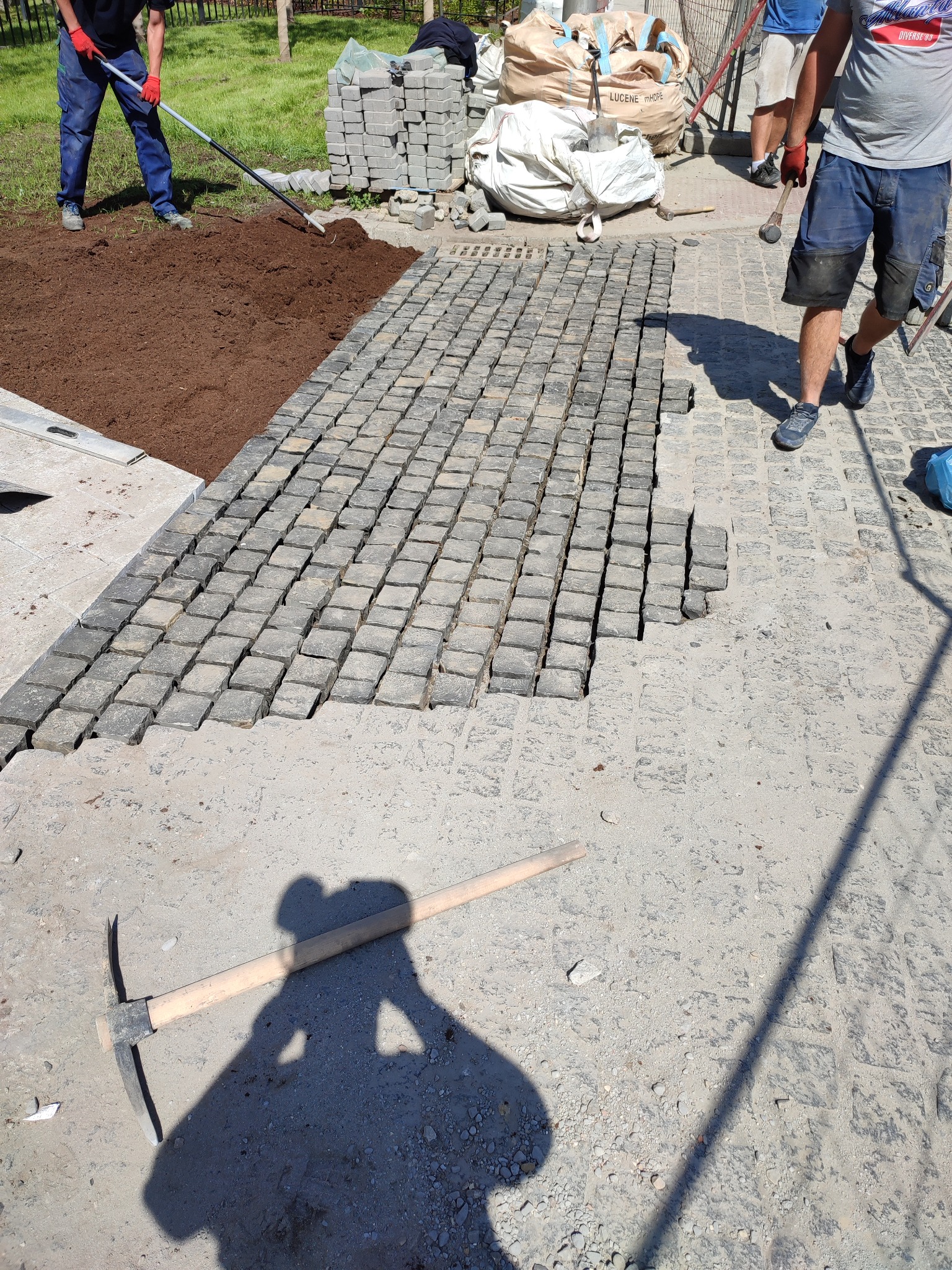 Fragment nawierzchni z kostki granitowej w trakcie układania; widoczne przygotowane podłoże, ułożona część nawierzchni, narzędzia i pracownicy.