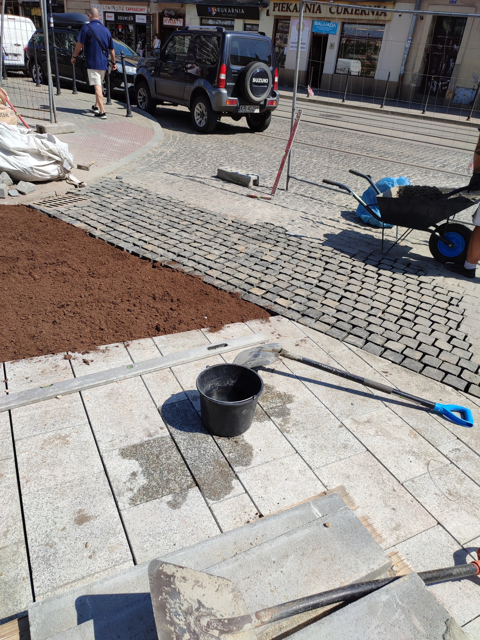 Układanie kostki brukowej na ulicy: widoczny fragment nowej nawierzchni z kostki granitowej, przygotowane podłoże z ziemi, narzędzia pracy (szpadel, wiadro), w tle ulica z przechodniami...