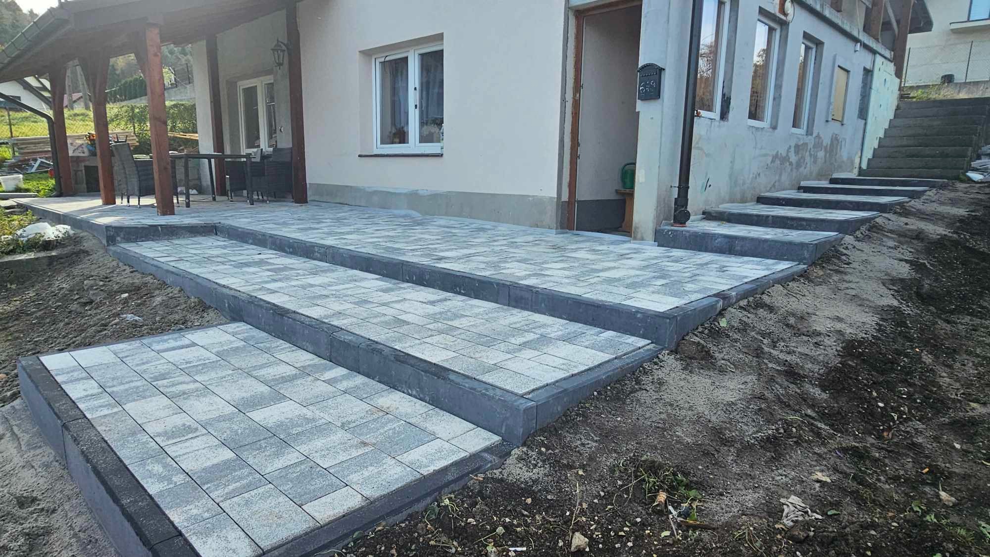Nowo ułożone schody z kostki brukowej prowadzące do domu z widoczną elewacją i elementami ogrodu w tle.