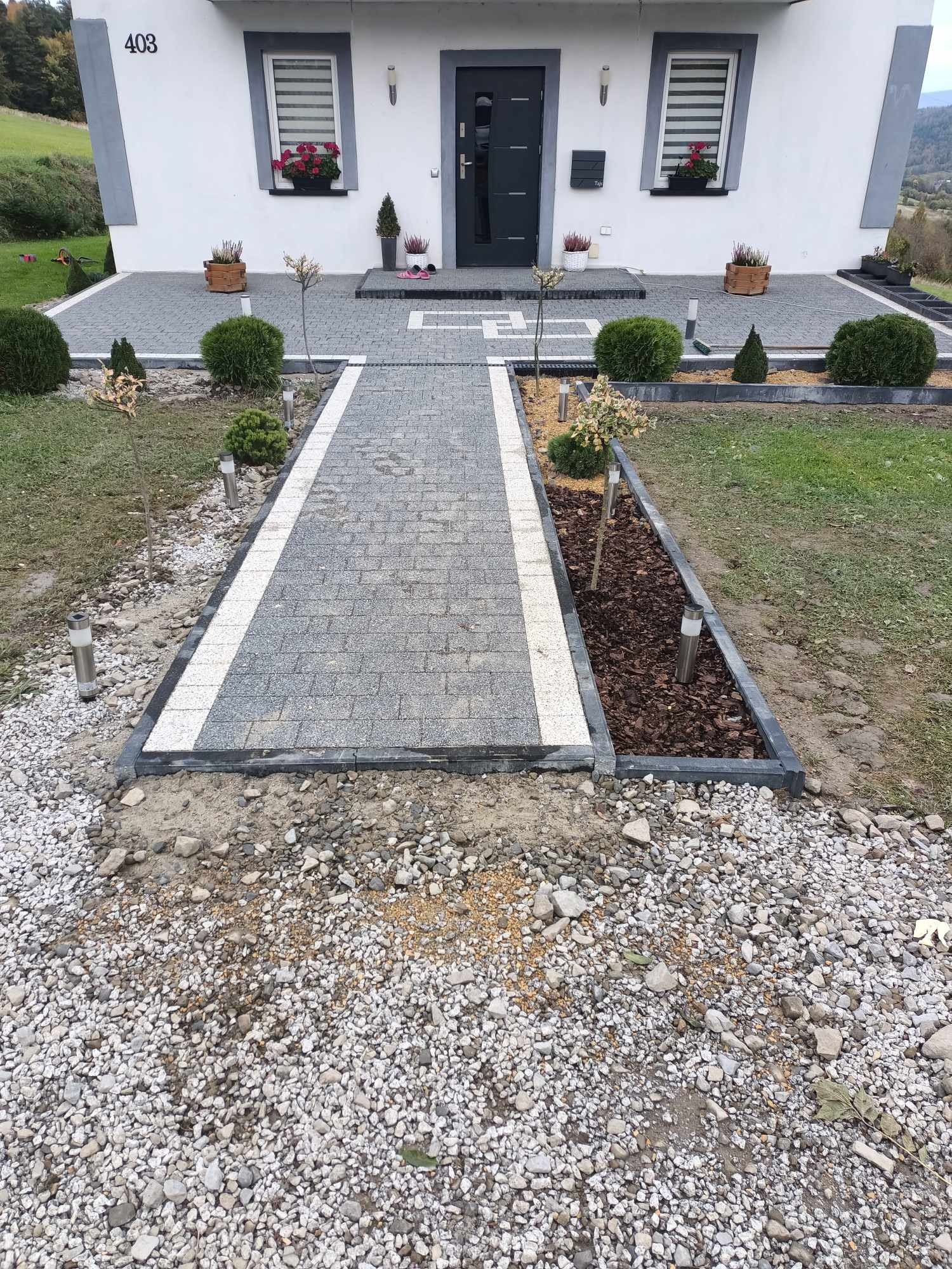 Chodnik z szarej kostki brukowej z białymi obrzeżami prowadzący do domu z numerem 403, obok rabaty z korą i niskimi roślinami, oświetlenie chodnikowe.
