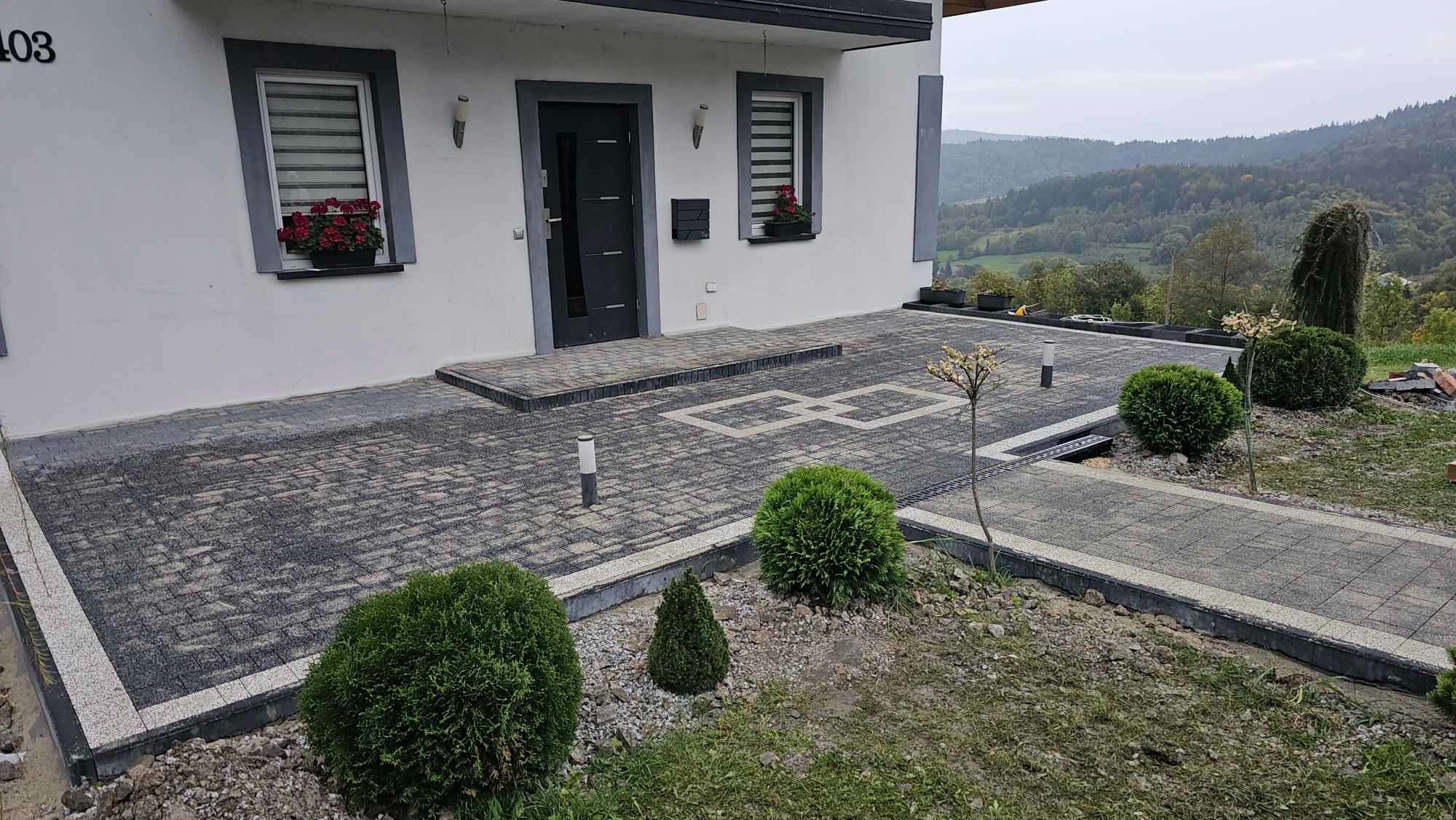 Nowoczesny podjazd z szarej kostki brukowej ułożonej we wzór, obrzeża z jasnej kostki, oświetlenie w postaci niskich lamp, zieleń ozdobna, w tle dom z szarą stolarką i widok na zalesione wzgórza.