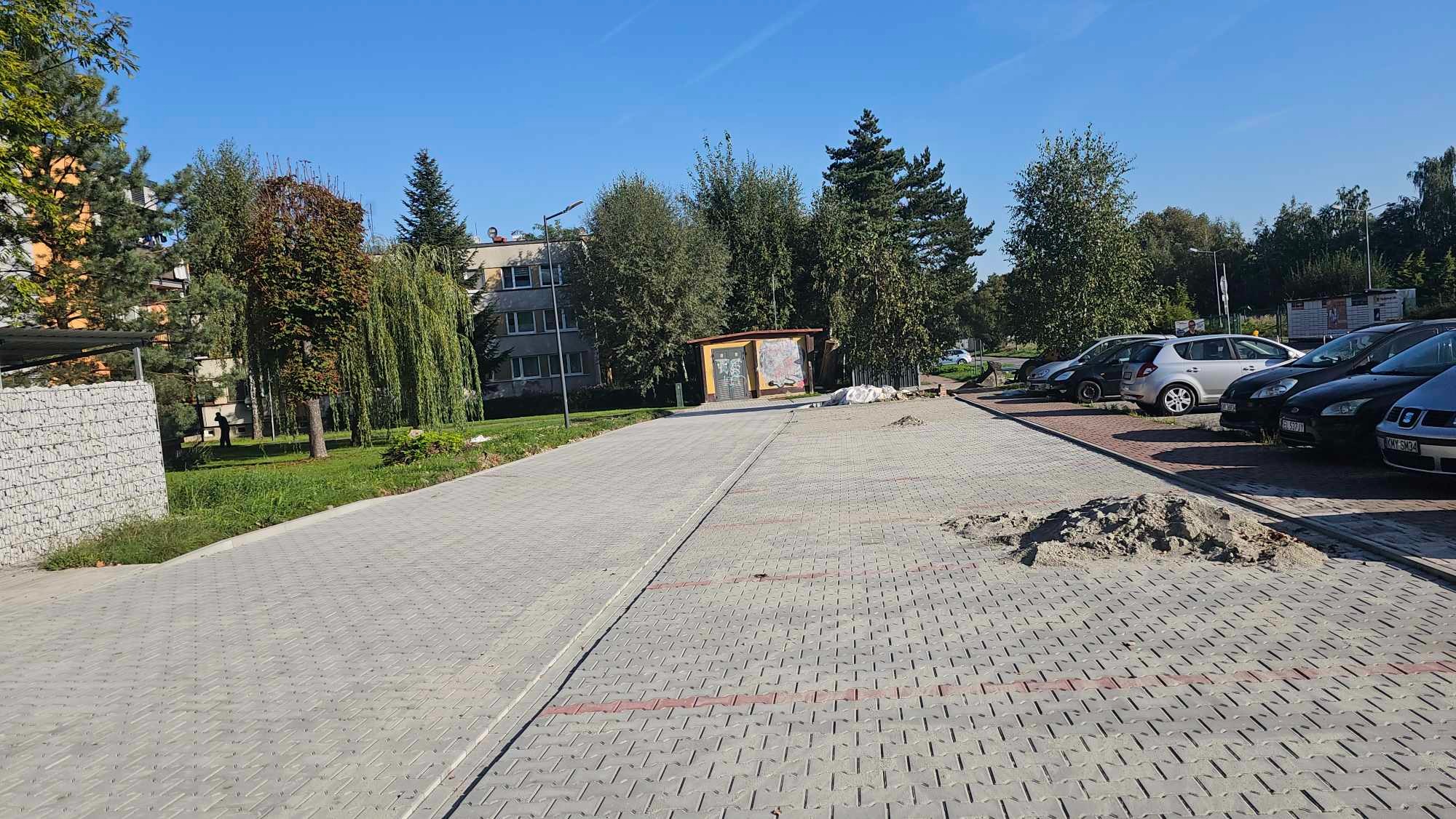 Szeroki kadr na nową nawierzchnię z szarej kostki brukowej na parkingu, z widocznymi śladami piasku i zaparkowanymi samochodami w tle.