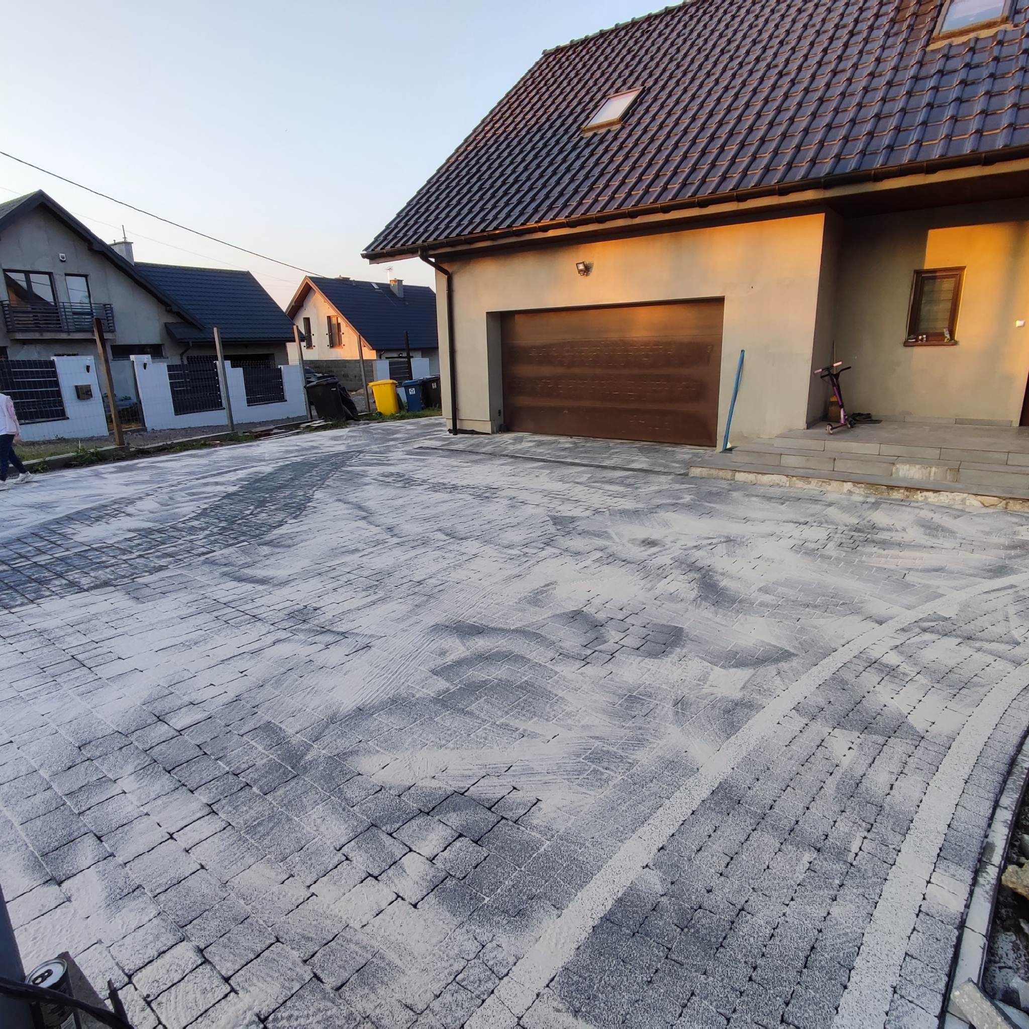Nowo ułożona, szara kostka brukowa na podjeździe do domu jednorodzinnego, zasypana piaskiem, z widocznymi śladami po oponach samochodu.