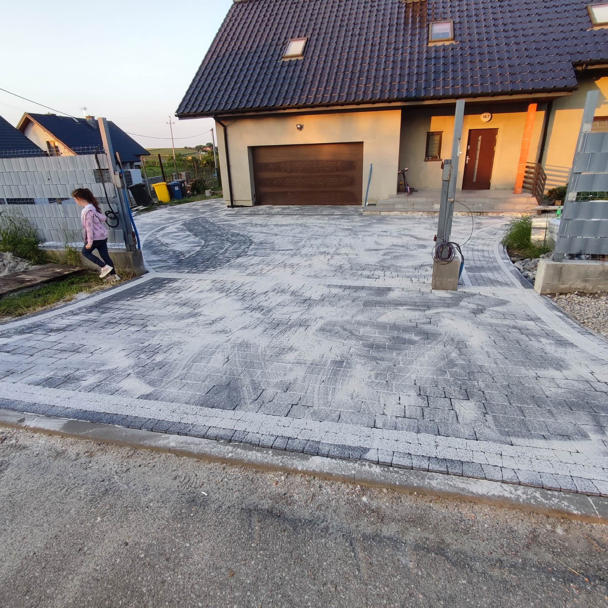 Nowo ułożona, szara kostka brukowa na podjeździe do domu jednorodzinnego, z widocznymi śladami piasku stabilizującego i przechodząca obok bramy wjazdowej, po której przechodzi dziewczynka.