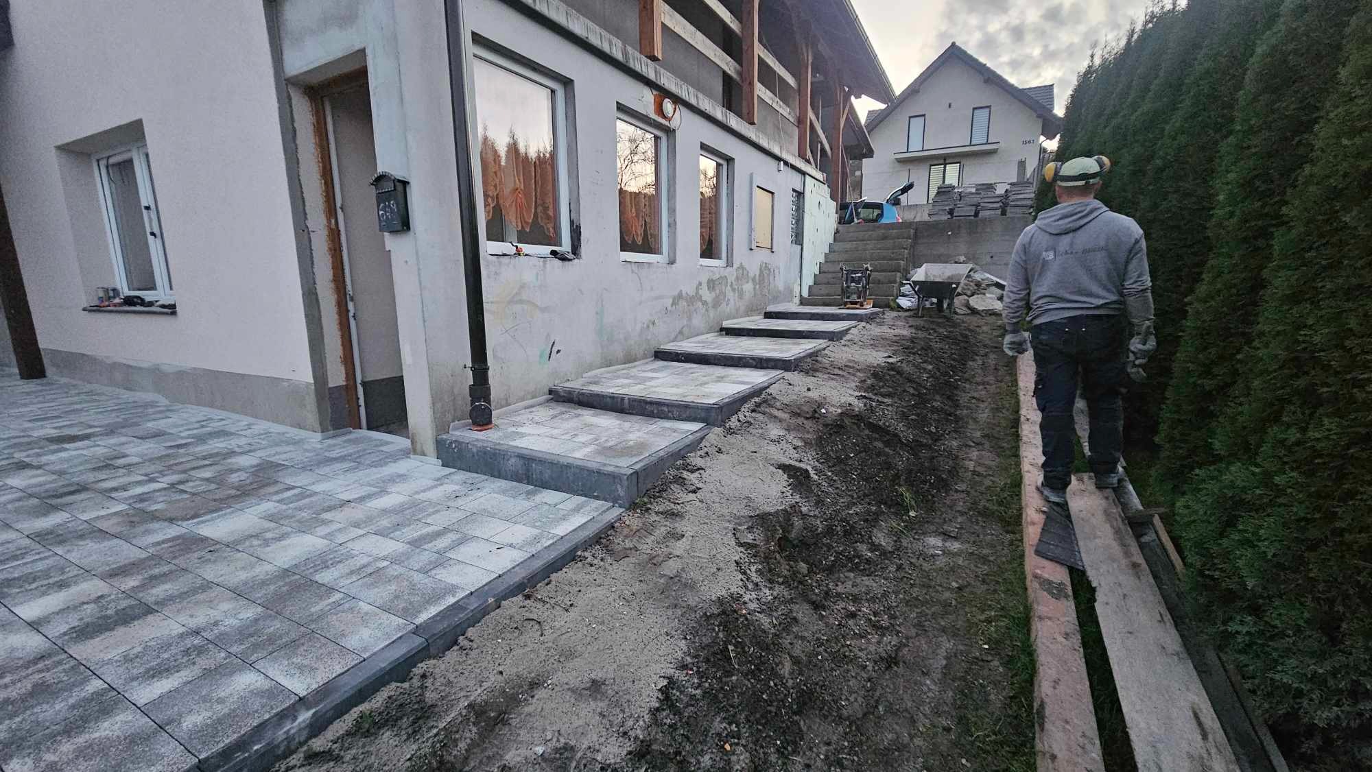 Układanie kostki brukowej w Tokarni: widok na ułożony fragment chodnika z kostki brukowej prowadzącego do budynku, w tle schody kamienne w trakcie budowy i pracownik na drewnianej kładce.