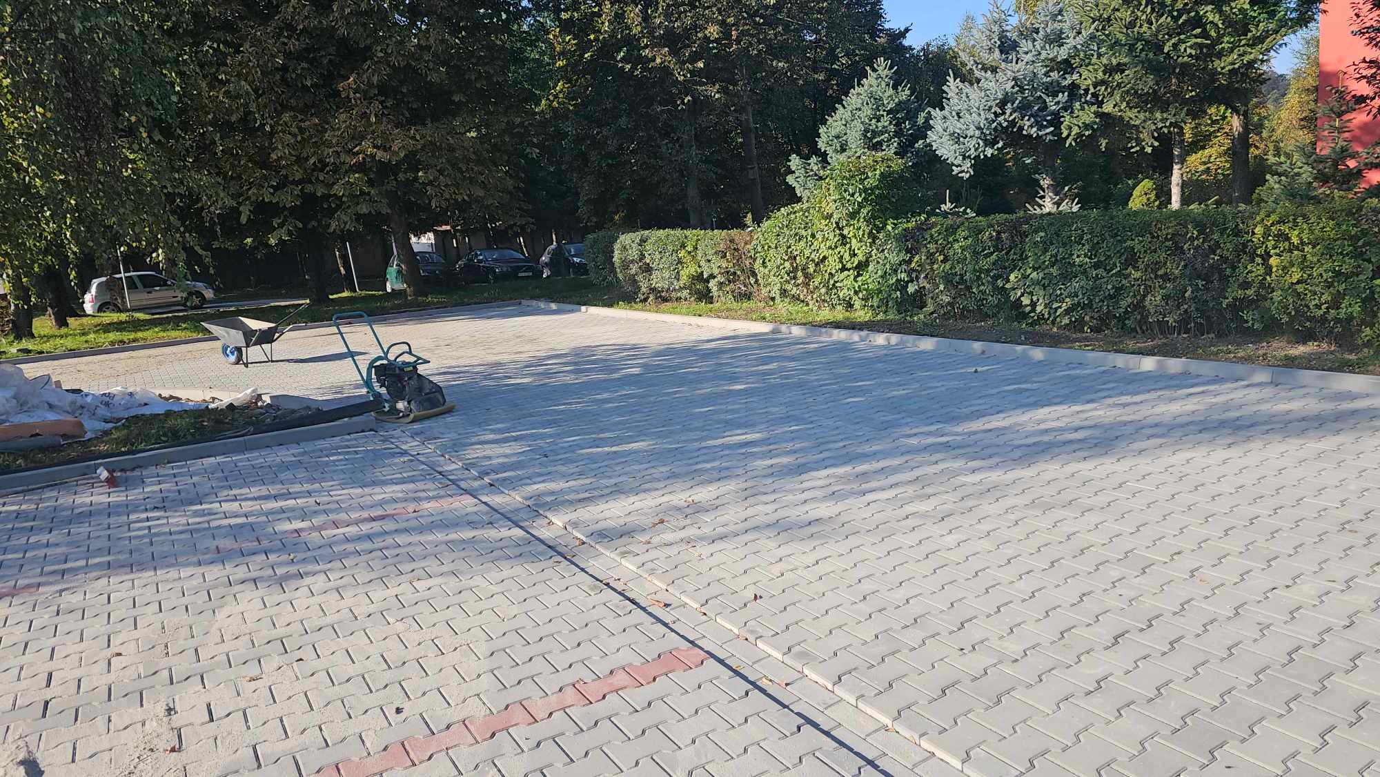 Ukończona nawierzchnia z szarej kostki brukowej typu Holland na parkingu, z widocznymi śladami piasku i ubijarką w tle, otoczona zielenią.