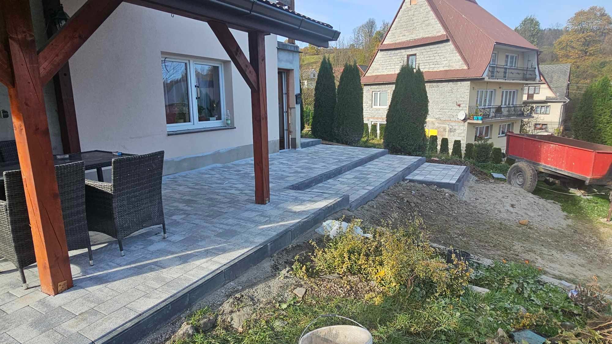 Nowoczesny taras z kostki brukowej w odcieniach szarości, z trzema schodami prowadzącymi do domu, widoczne meble ogrodowe i częściowo przygotowany teren wokół.