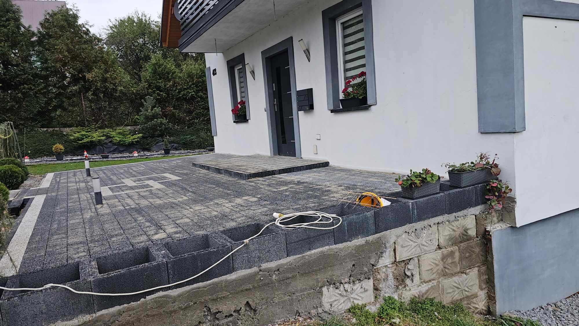 Ułożona kostka brukowa przed domem z widocznymi elementami podwyższenia terenu z betonowych bloczków i kablem zasilającym narzędzie.
