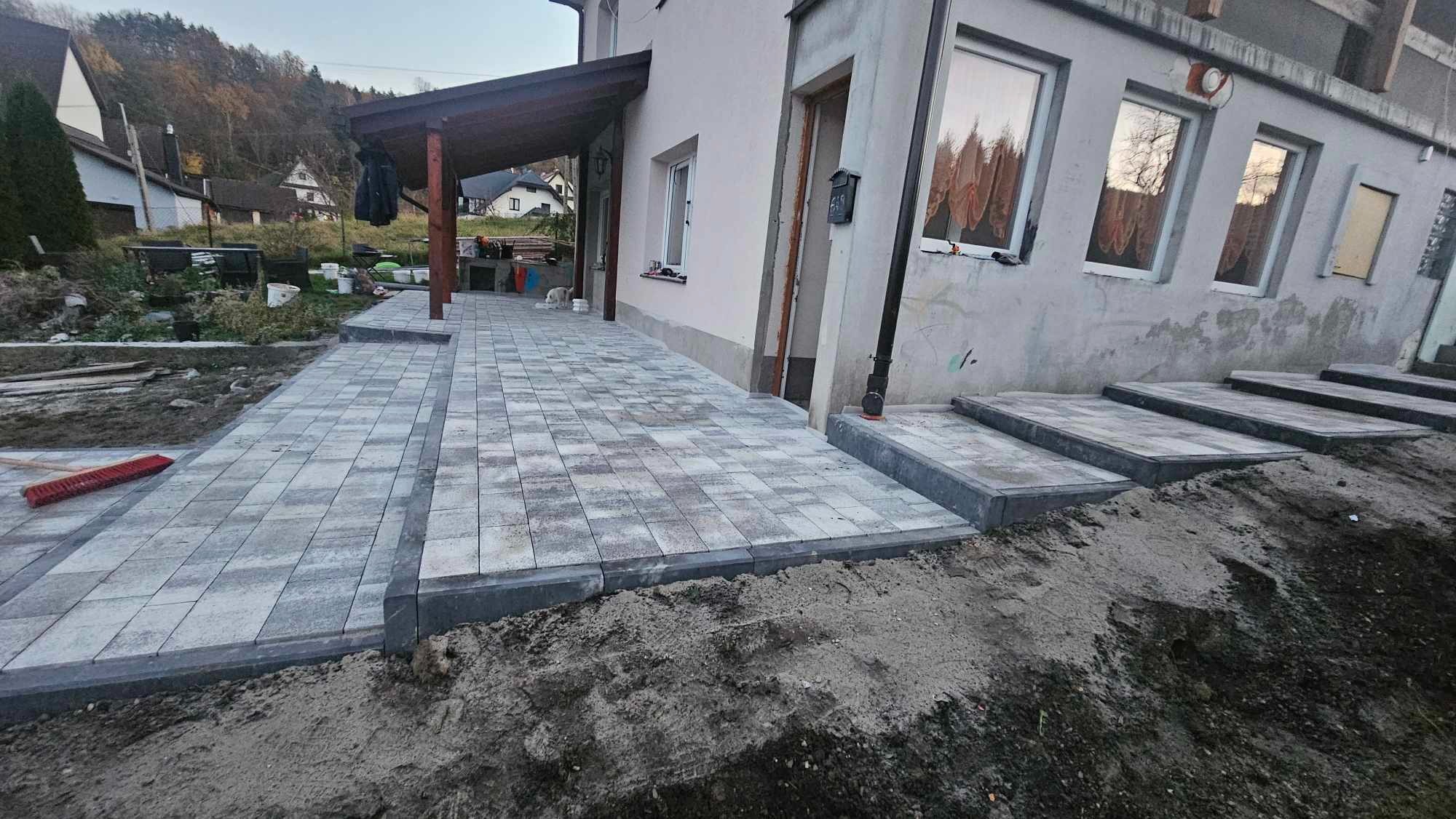 Nowo ułożona, szara kostka brukowa przed domem z widocznymi schodami wejściowymi i częściowo zagospodarowanym terenem wokół.