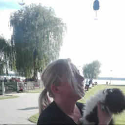 Kobieta z blond włosami trzymająca małego czarno-białego psa na tle parku z jeziorem i wierzbami płaczącymi.