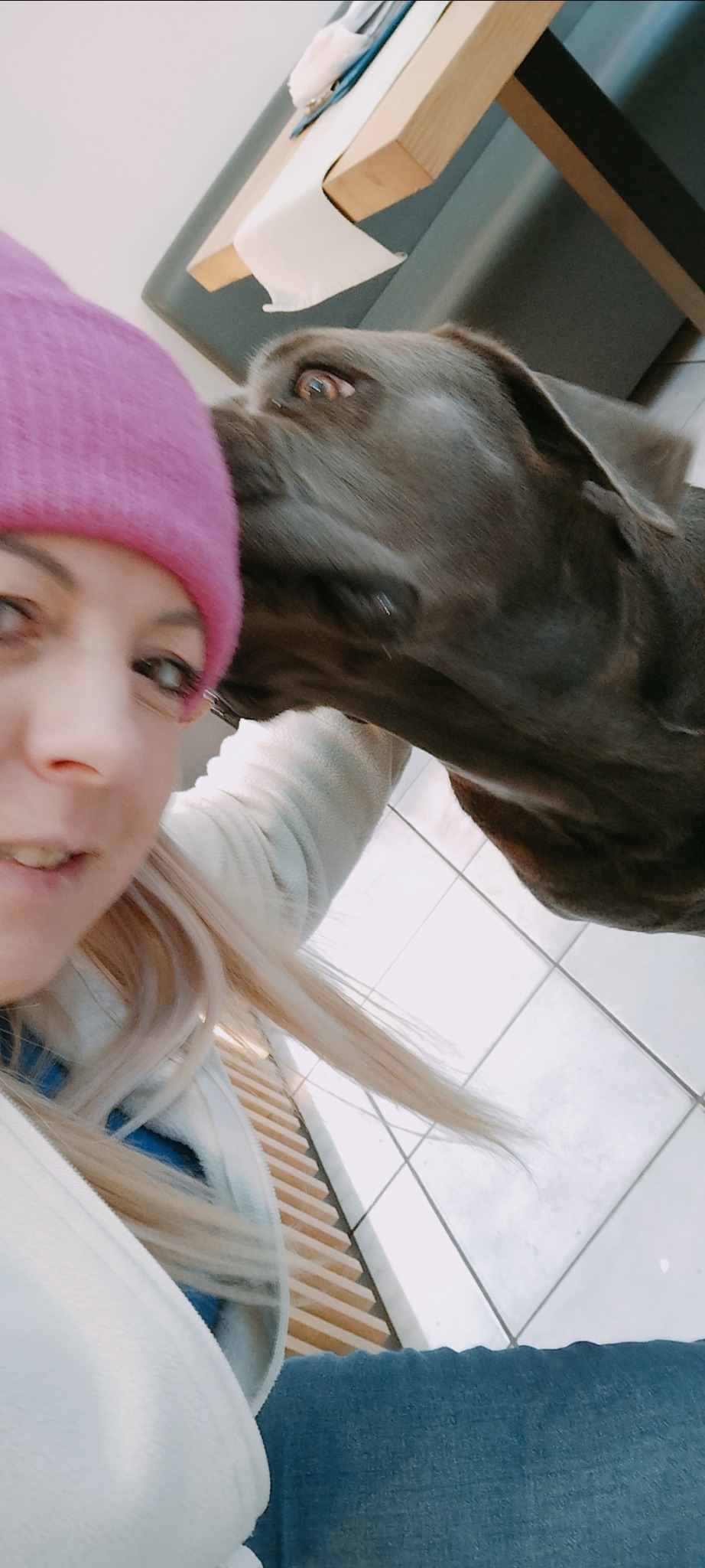 Kobieta w różowej czapce robi selfie z dużym, szarym psem, który liże ją po twarzy w jasnym wnętrzu z kafelkami na podłodze i drewnianym elementem w tle.