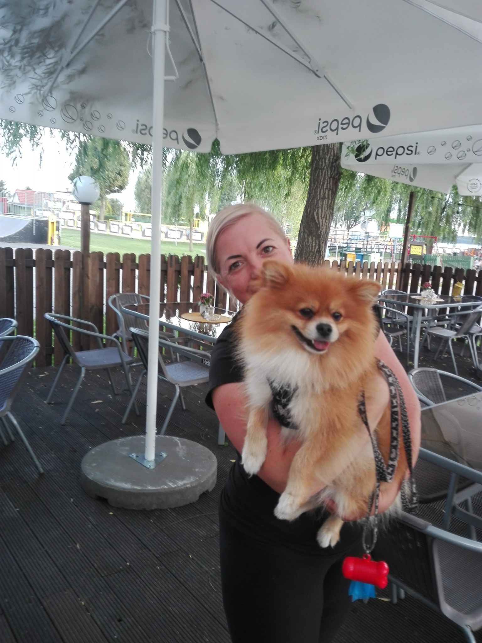 Uśmiechnięta kobieta trzyma na rękach rudego szpica miniaturowego z kolorową zabawką na smyczy, siedząc na tarasie kawiarni z parasolami Pepsi w tle.