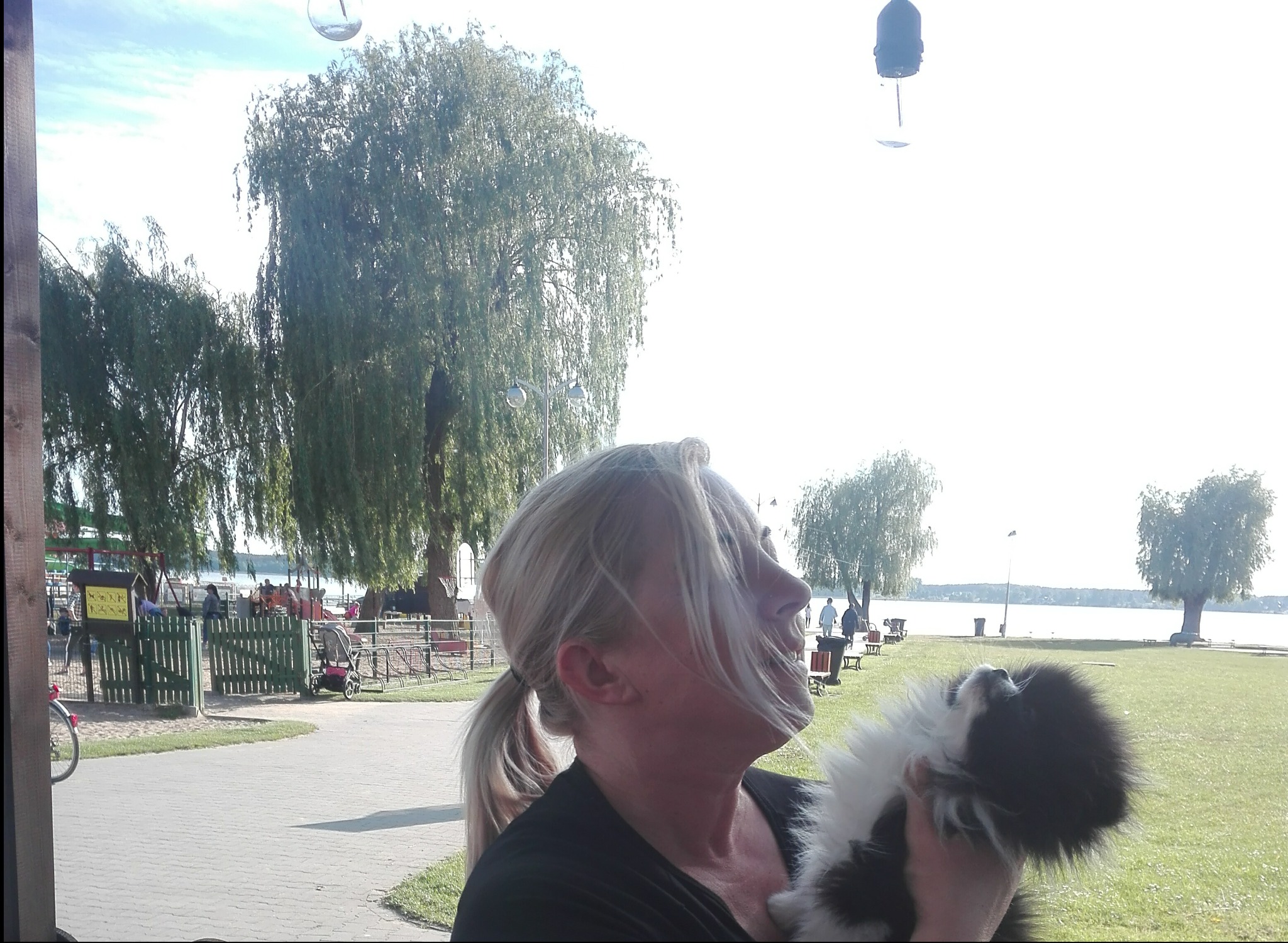 Kobieta z blond włosami trzymająca małego czarno-białego psa na tle parku z jeziorem i wierzbami płaczącymi.