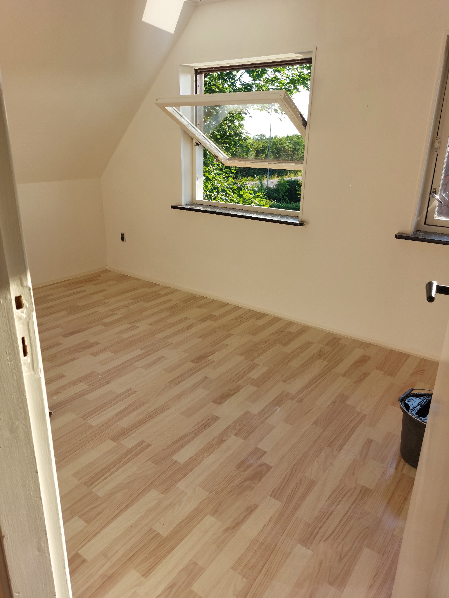 Pomalowane na biało poddasze z drewnianą podłogą, otwartym oknem i widokiem na zieleń, z wiaderkiem z farbą w rogu.
