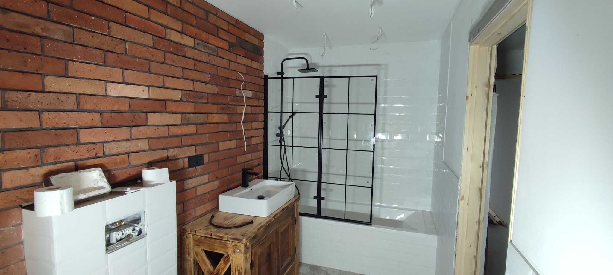 Łazienka w trakcie wykańczania: ściana z czerwonej cegły, biała umywalka na drewnianej szafce, czarna kabina prysznicowa z szybami w kratkę, białe płytki na ścianach i niedokończona zabudowa toalety.