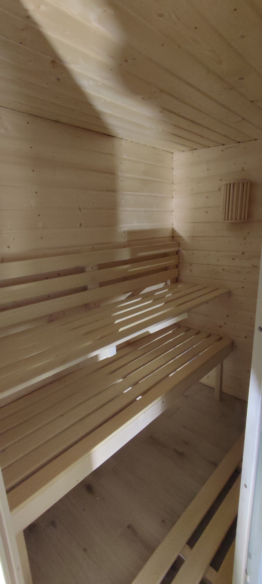 Wnętrze sauny z drewnianymi ławkami na dwóch poziomach, drewnianymi ścianami i podłogą z paneli, widoczne źródło światła na ścianie.