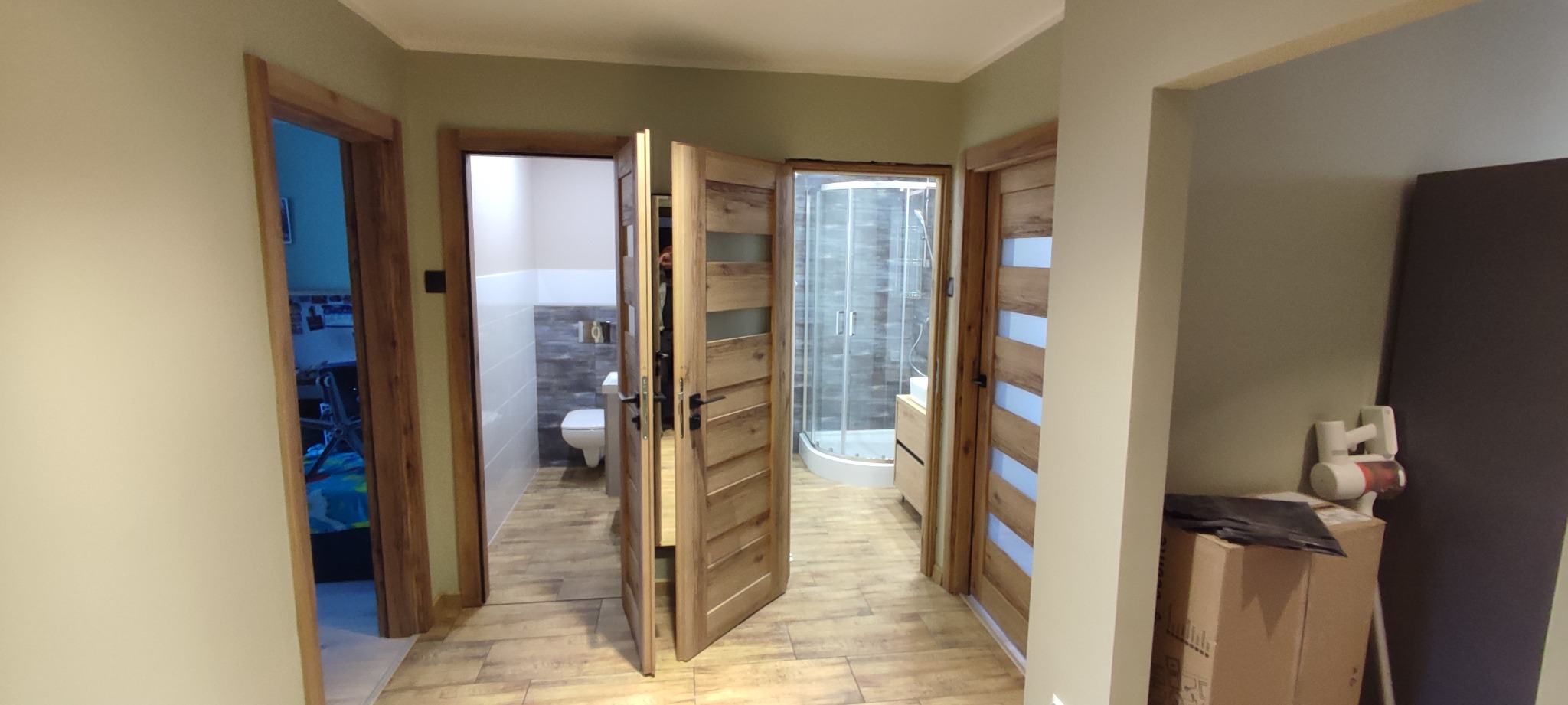 Korytarz z trzema otwartymi drzwiami z drewnianymi ościeżnicami i panelami z mlecznego szkła, prowadzącymi do łazienki z toaletą, łazienki z kabiną prysznicową i pokoju z niebieskim łóżkiem,...