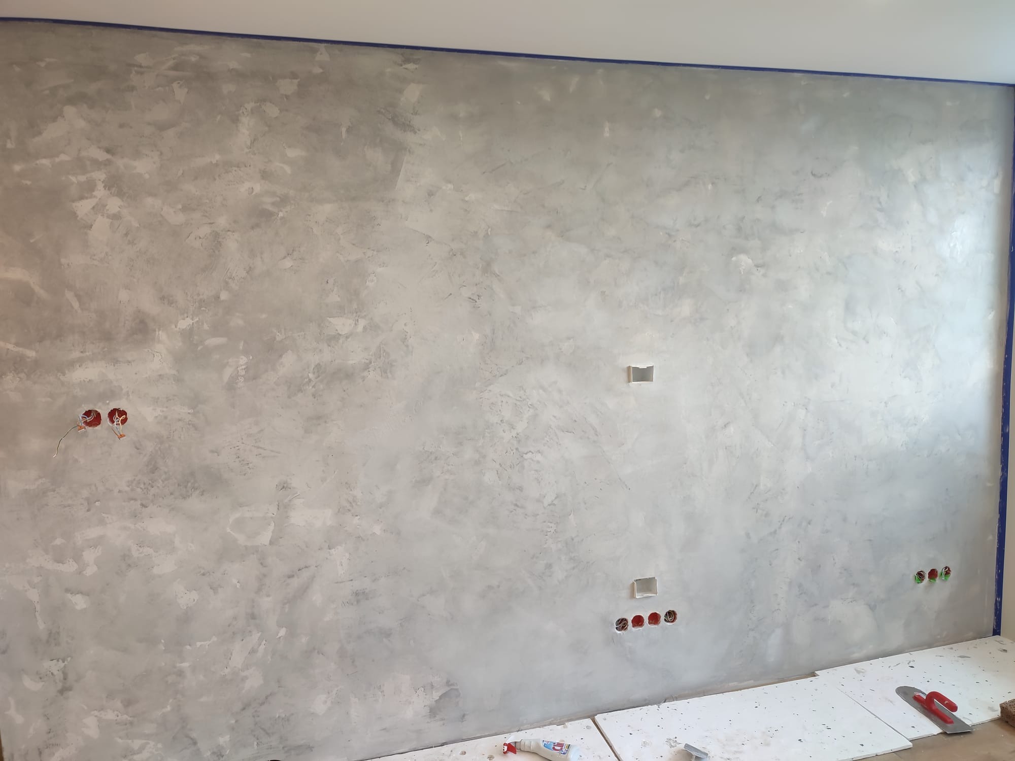 Ściana z efektem dekoracyjnym w odcieniach szarości, zabezpieczona niebieską taśmą malarską przy suficie i podłodze, widoczne puszki elektryczne i narzędzia malarskie na podłodze.