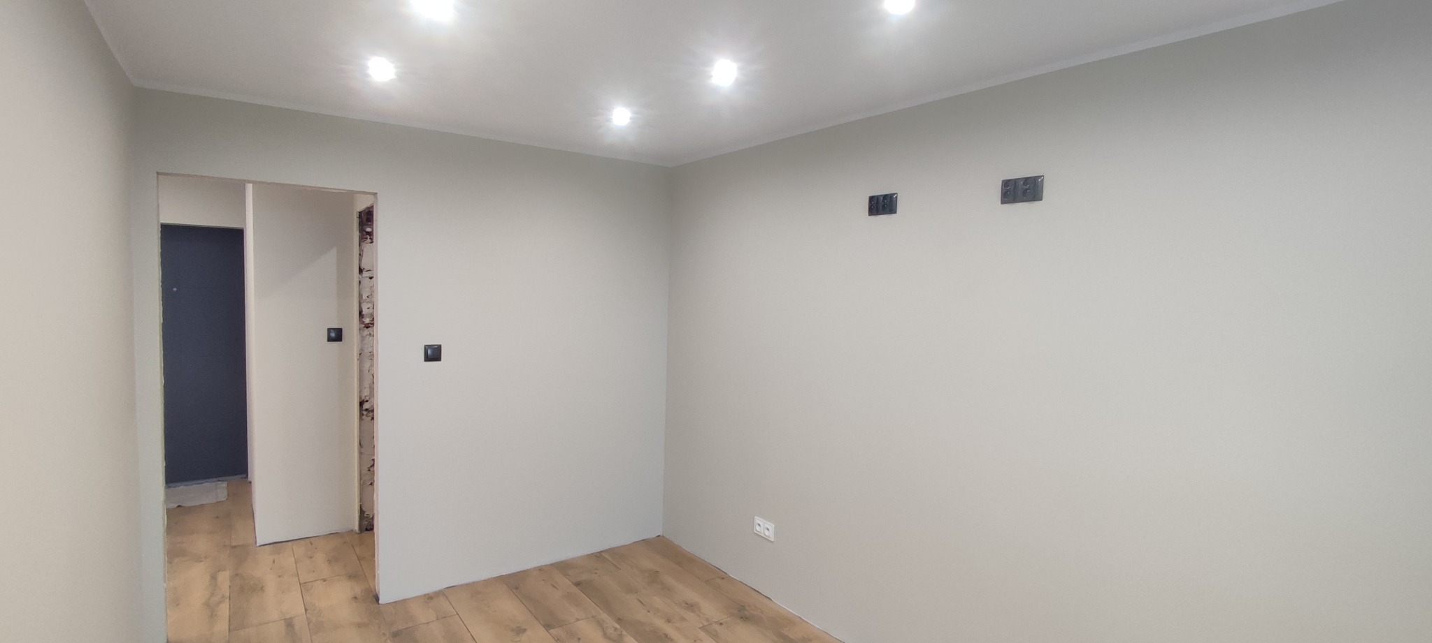 Pokój po świeżym malowaniu na szaro, z drewnopodobną podłogą, widoczne gniazdka elektryczne i otwory na kable, sufit z wbudowanym oświetleniem punktowym, drzwi bez ościeżnicy w trakcie montażu.