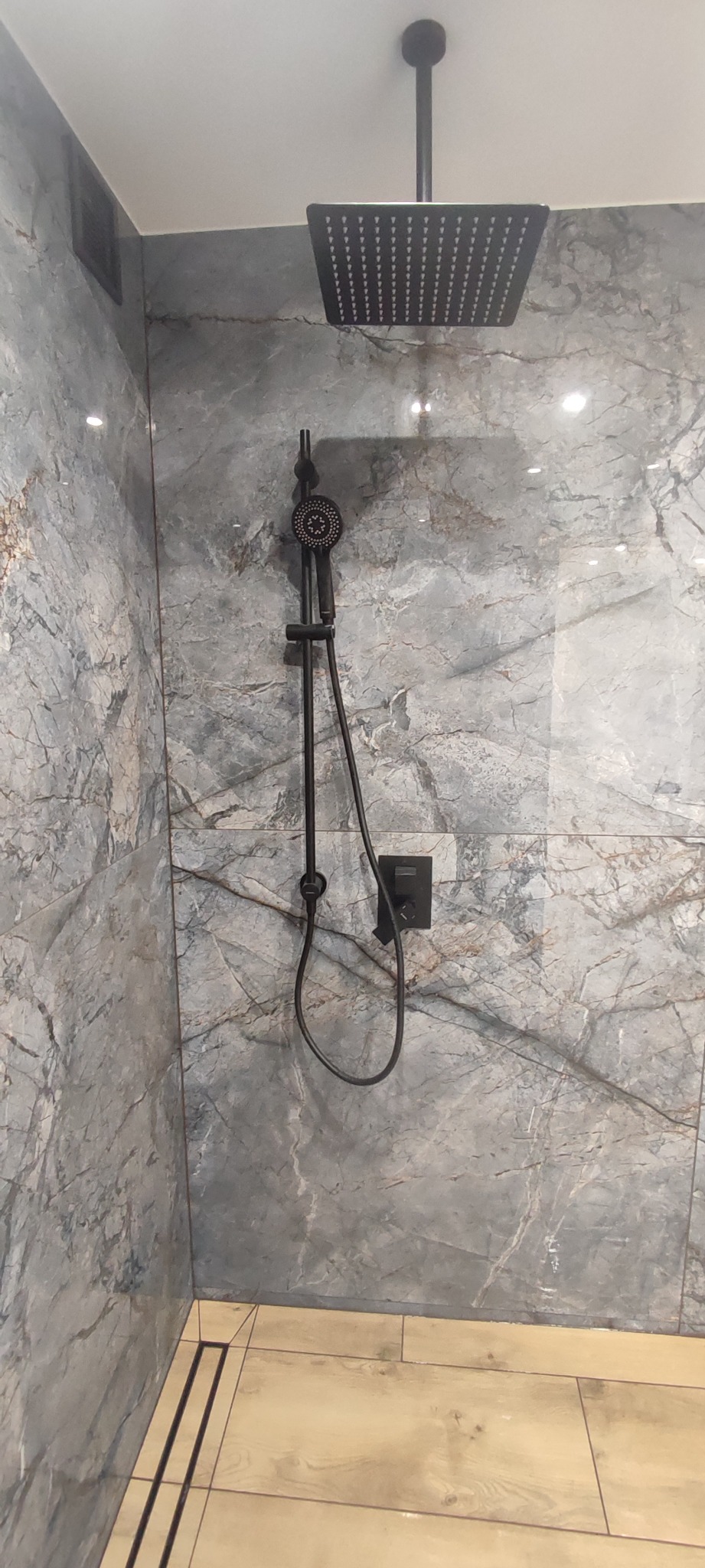 Nowoczesna kabina prysznicowa z czarną deszczownicą i słuchawką na tle marmurowych płytek w odcieniach szarości i brązu, z widocznym odpływem liniowym w podłodze.