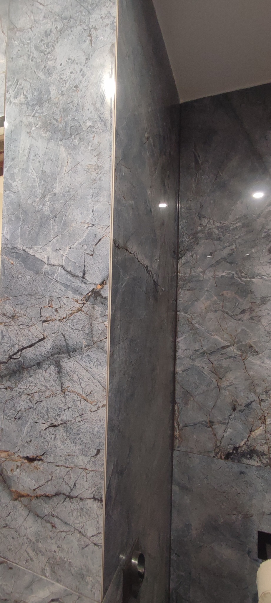 Ściana wyłożona szarymi płytami imitującymi marmur z wyraźnym żyłkowaniem, narożnik pomieszczenia, widoczne oświetlenie sufitowe.