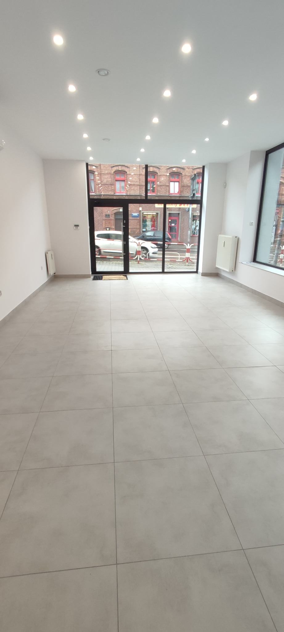 Wnętrze nowo wykończonego lokalu usługowego z szarą, kwadratową podłogą z płytek, białymi ścianami i punktowym oświetleniem sufitowym, widok na ulicę przez przeszklone drzwi wejściowe.