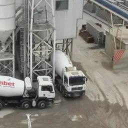 Trzy betoniarki firmy Rembet na placu budowy z silosami i elementami prefabrykowanymi w tle, widok z góry.