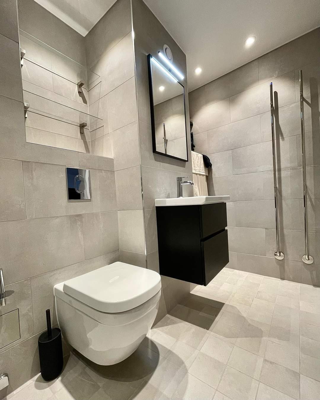 Nowoczesna łazienka z szarymi płytkami, podwieszaną toaletą, czarną szafką pod umywalką i podświetlanym lustrem, minimalistyczny design z chromowanymi akcentami.