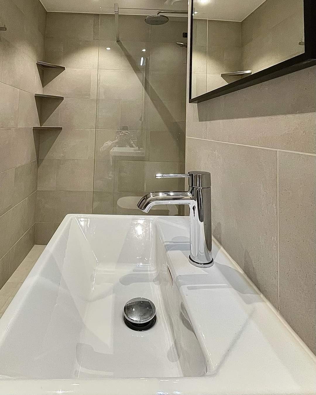 Nowoczesna łazienka z kwadratową umywalką, chromowaną baterią i szklaną kabiną prysznicową, podświetlone półki narożne na tle płytek w odcieniach szarości.