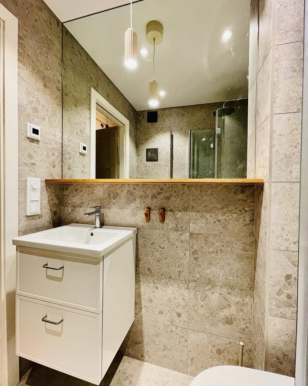 Nowoczesna łazienka z szarymi marmurkowymi płytkami, białą szafką pod umywalkę z dwiema szufladami i dużym lustrem odbijającym kabinę prysznicową. Nad umywalką drewniana półka i dwa drewniane haczyki.