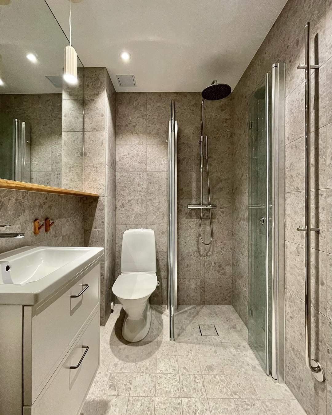 Nowoczesna łazienka z szarymi płytkami imitującymi marmur, biała umywalka z szafką i toaleta, kabina prysznicowa z deszczownicą, minimalistyczne oświetlenie.