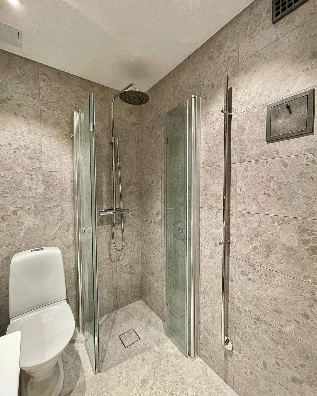 Nowoczesna łazienka wykończona szarymi płytkami imitującymi marmur, z kabiną prysznicową z transparentnego szkła, białą toaletą i grzejnikiem drabinkowym.