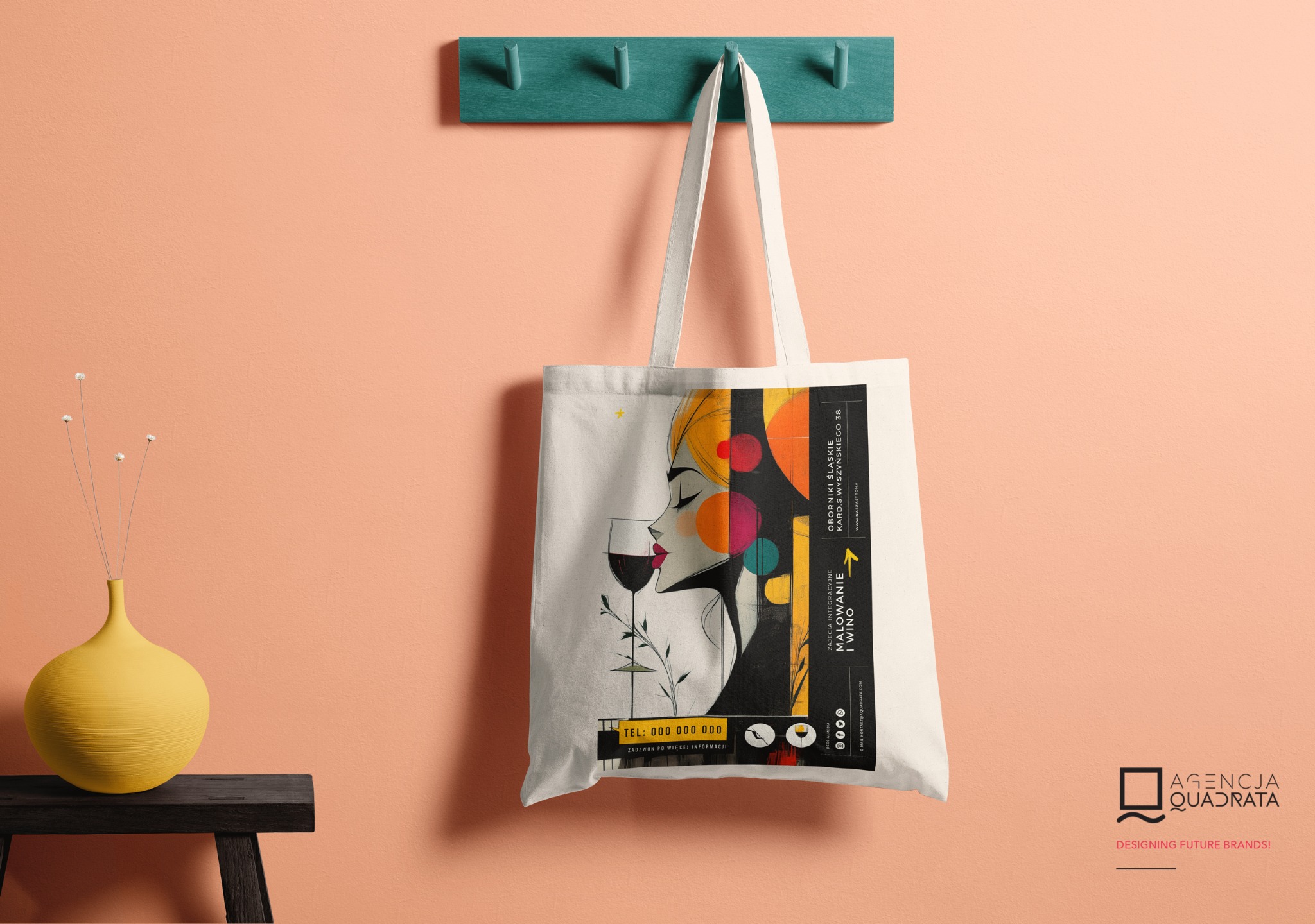 Ekologiczna torba z nadrukiem graficznym kobiety pijącej wino, zawieszona na wieszaku ściennym w minimalistycznym wnętrzu z żółtym wazonem i ciemnym stolikiem.