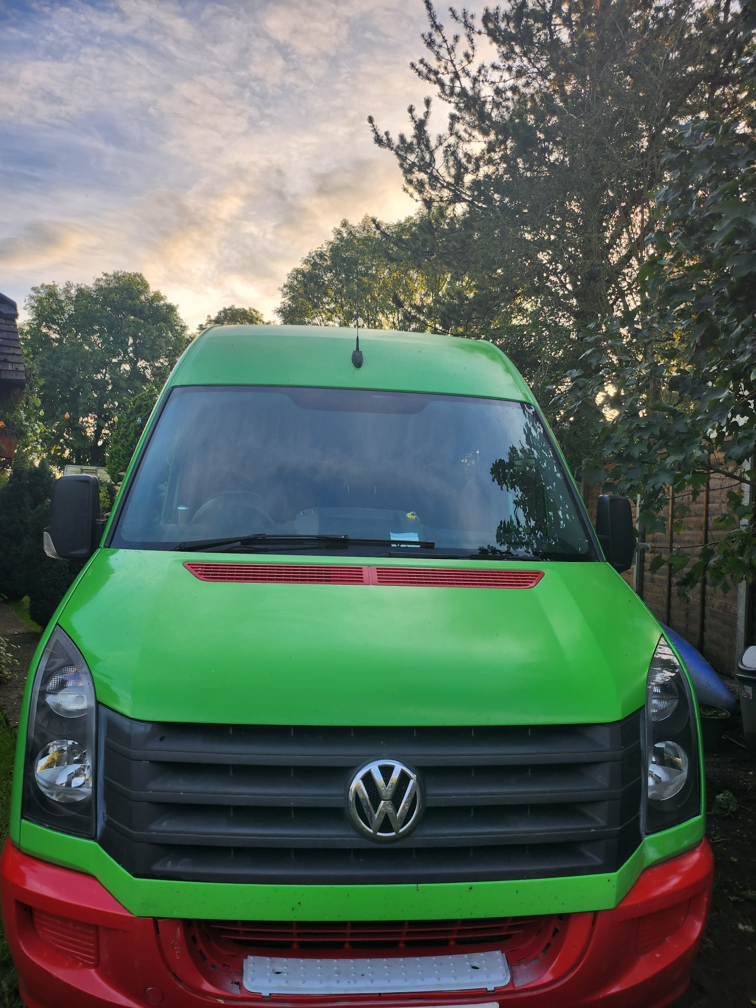 Zielono-czerwony Volkswagen Crafter zaparkowany na podwórku, widok z przodu na tle drzew i nieba o zmierzchu.