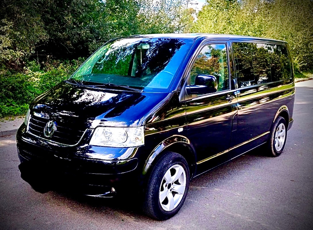 Czarny, lśniący Volkswagen Transporter T5 na asfaltowej drodze, otoczony zielenią drzew, z białymi felgami i widocznym logo VW.