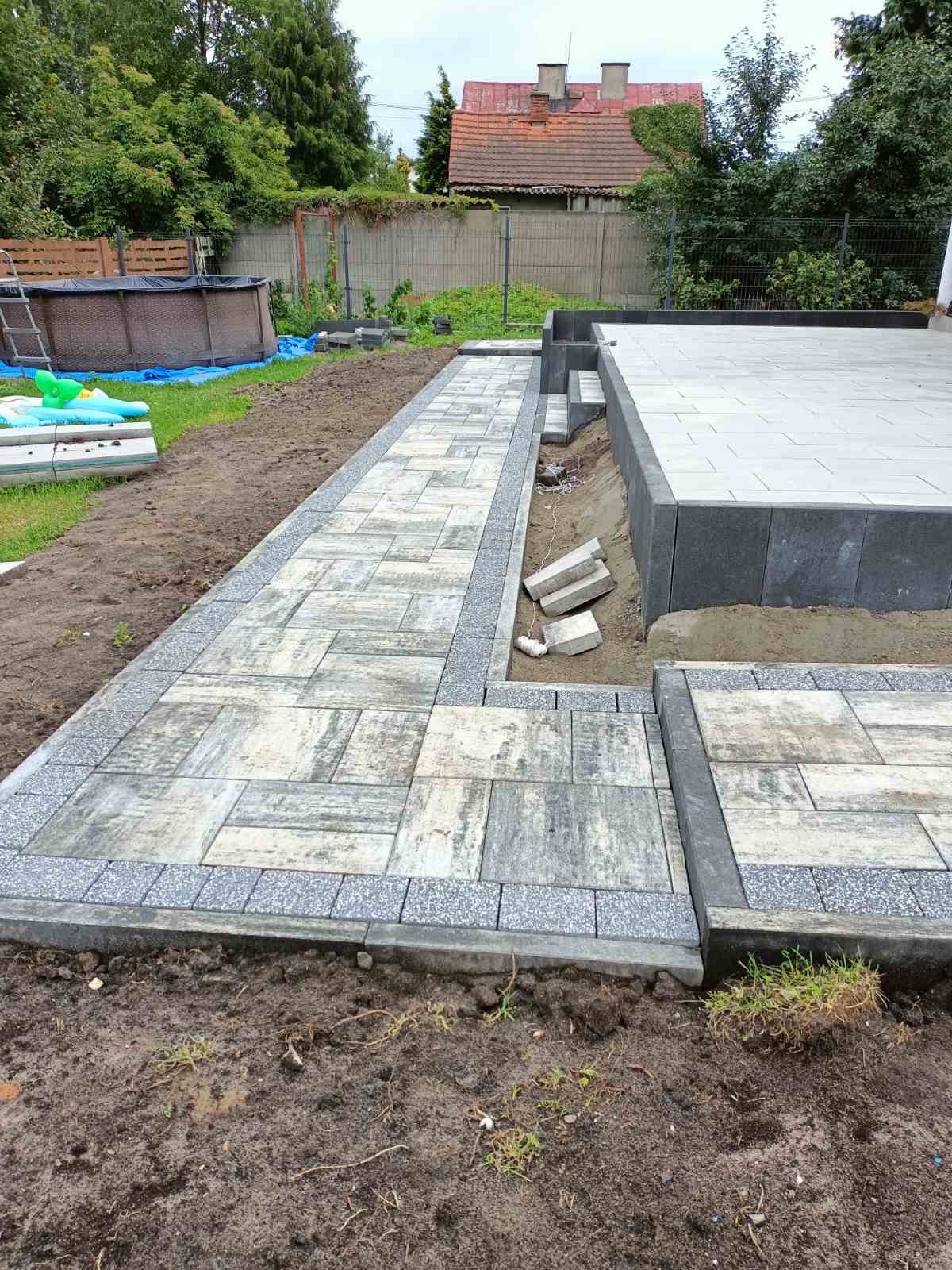 Świeżo ułożona ścieżka z kostki brukowej w odcieniach szarości i beżu, obramowana drobną kostką granitową, prowadząca do podwyższonego tarasu z szarych płyt; w tle widoczny basen ogrodowy i dom...