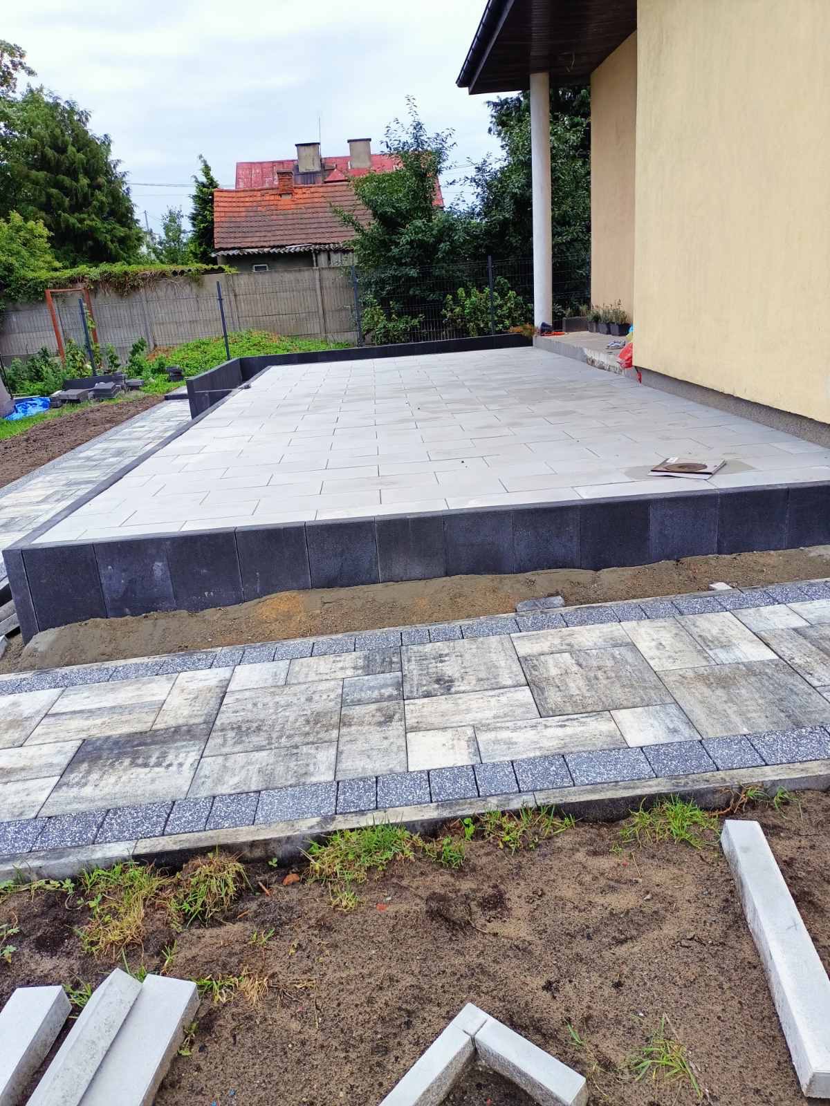 Nierówny teren z częściowo ułożonym chodnikiem z szarej kostki brukowej, obrzeża z ciemnych elementów, widoczne niedokończone prace i przygotowane elementy do ułożenia.