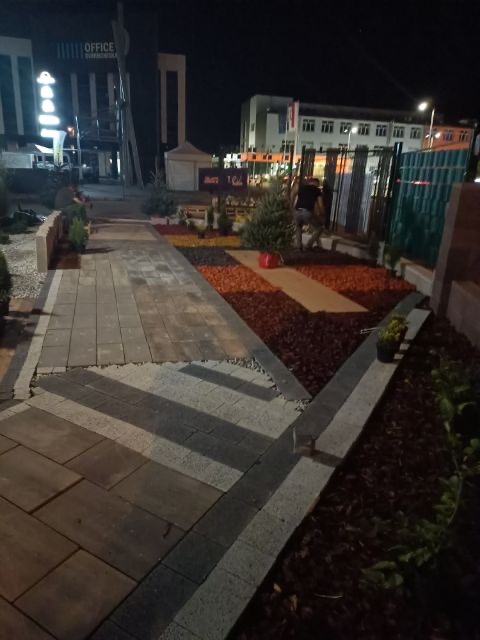 Nocne ujęcie ścieżki z kostki brukowej ułożonej we wzór szewronu, obrzeża z kostki granitowej, rabaty z korą i roślinami iglastymi, w tle budynki biurowe i oświetlenie uliczne.