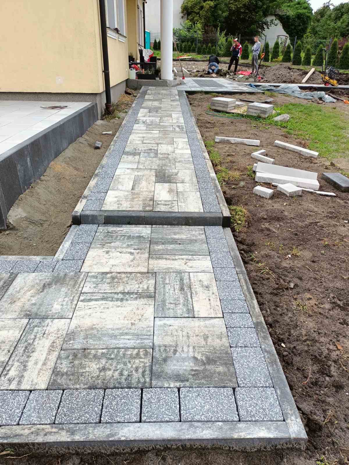 Ścieżka z kostki brukowej o nieregularnym wzorze, wykończona obrzeżem z kostki granitowej, prowadząca do domu we Włocławku. W tle widoczni pracownicy podczas dalszych prac brukarskich.