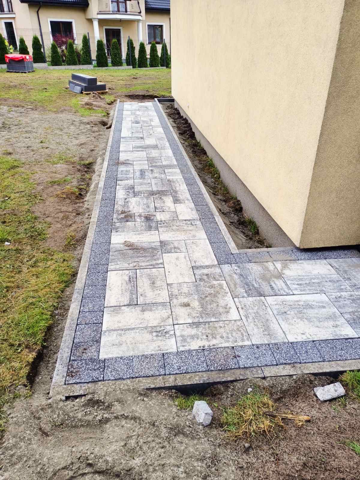 Ułożony chodnik z szarej kostki brukowej z obramowaniem z ciemniejszej kostki granitowej, prowadzący wzdłuż ściany budynku na trawnik z młodymi tujami w tle.