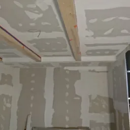 Wnętrze w trakcie prac wykończeniowych: ściany i sufit z płyt kartonowo-gipsowych, częściowo pokryte masą szpachlową, widoczne drewniane belki z pomarańczowymi elementami mocującymi na suficie...