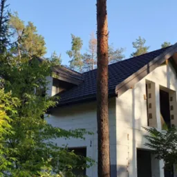 Dom w trakcie budowy z ciemnym dachem z blachodachówki, widoczny przez drzewa, w porannym słońcu.