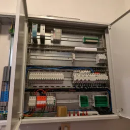 Otwarta szafa elektryczna z widocznymi zabezpieczeniami, przewodami, listwami zaciskowymi i innymi elementami instalacji elektrycznej.