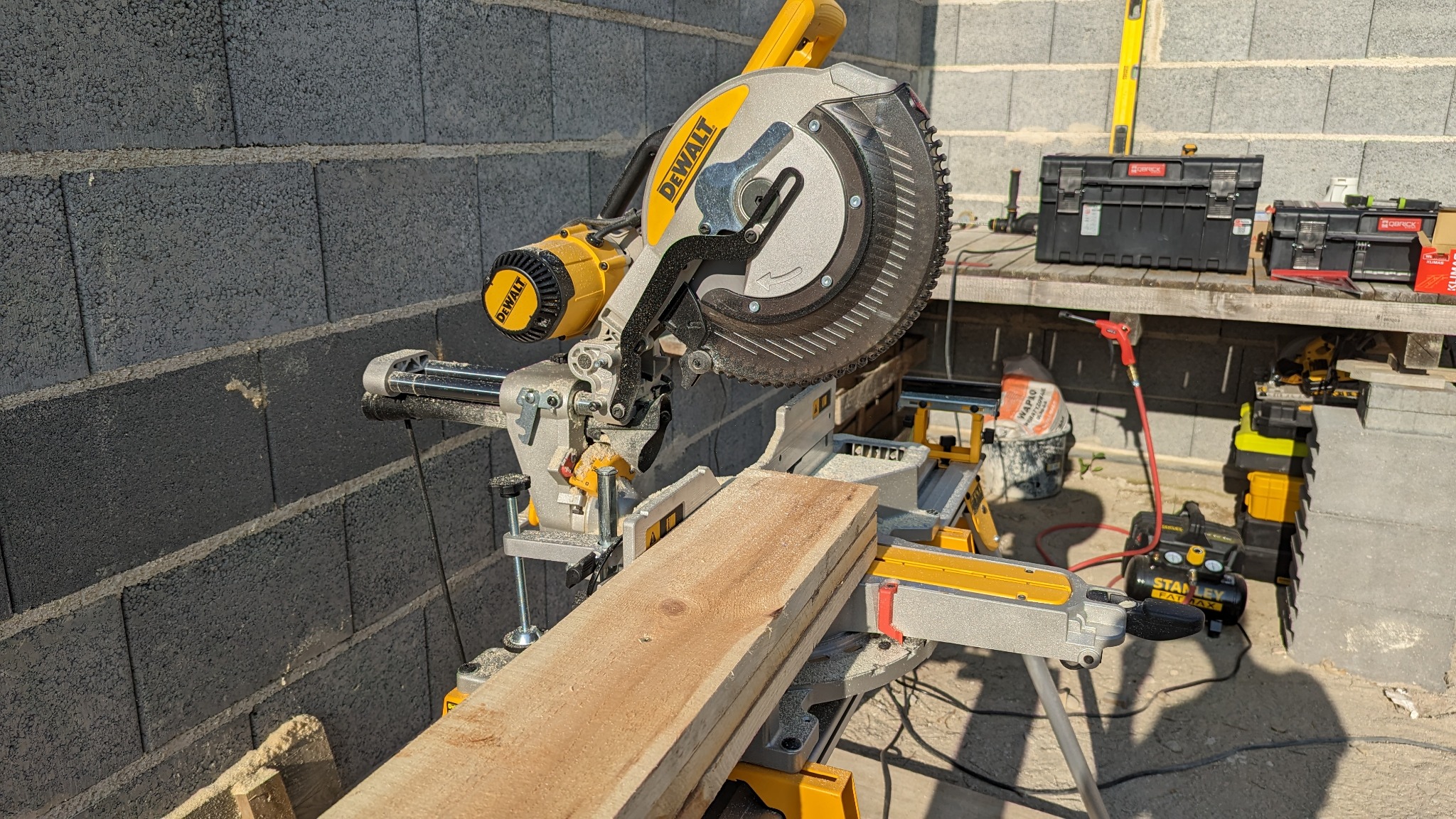 Piła ukośnica DeWalt przygotowana do cięcia deski na tle ściany z bloczków betonowych i narzędzi na stole warsztatowym.