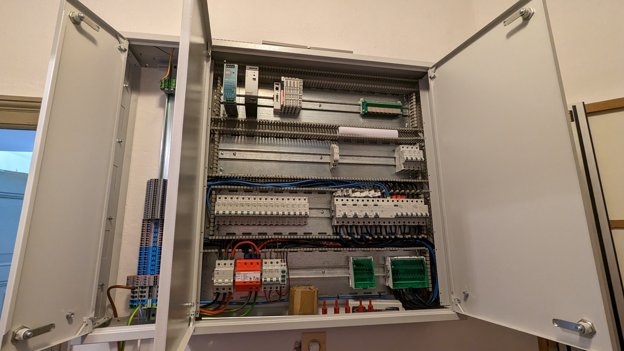 Otwarta szafa elektryczna z widocznymi zabezpieczeniami, przewodami, listwami zaciskowymi i innymi elementami instalacji elektrycznej.