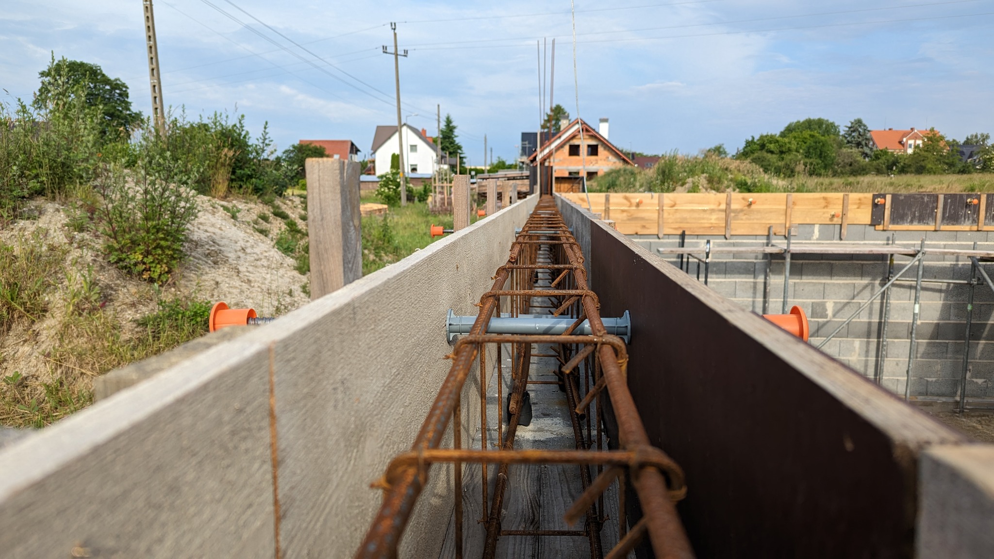 Zbrojenie fundamentów w szalunku, widok z wnętrza, przygotowane do zalania betonem na budowie domu w Bolesławcu, w tle dom w stanie surowym otwartym.